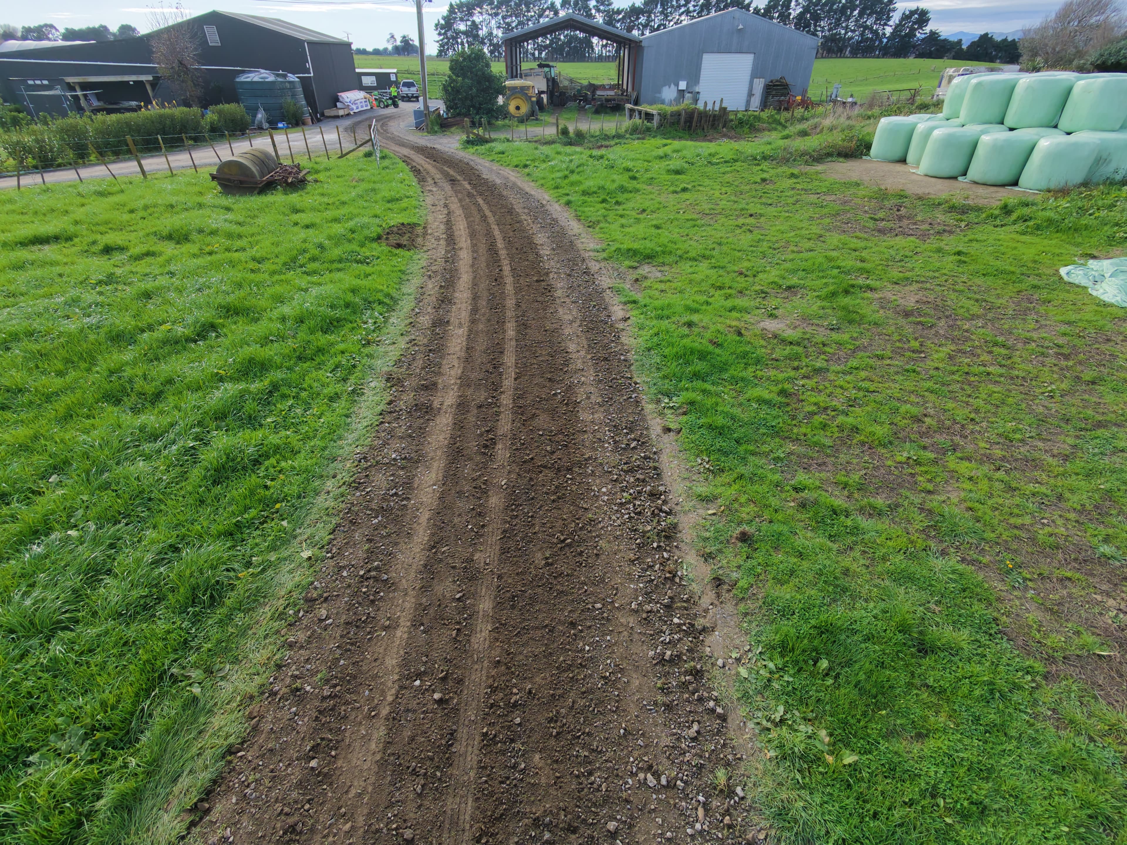 Curved dirt farm road through green fields leading to sheds and plastic-wrapped hay bales.